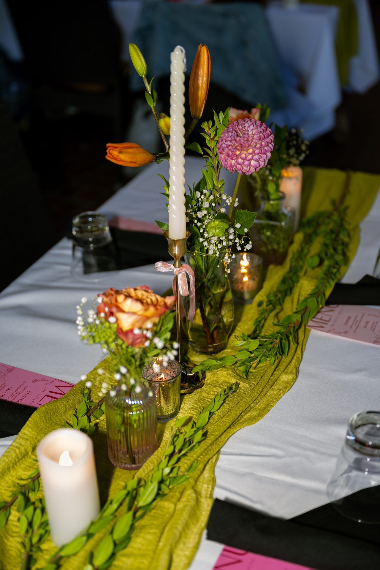 Coral and purple flowers, taper candles, and leafy branches glow along the olive table runner.