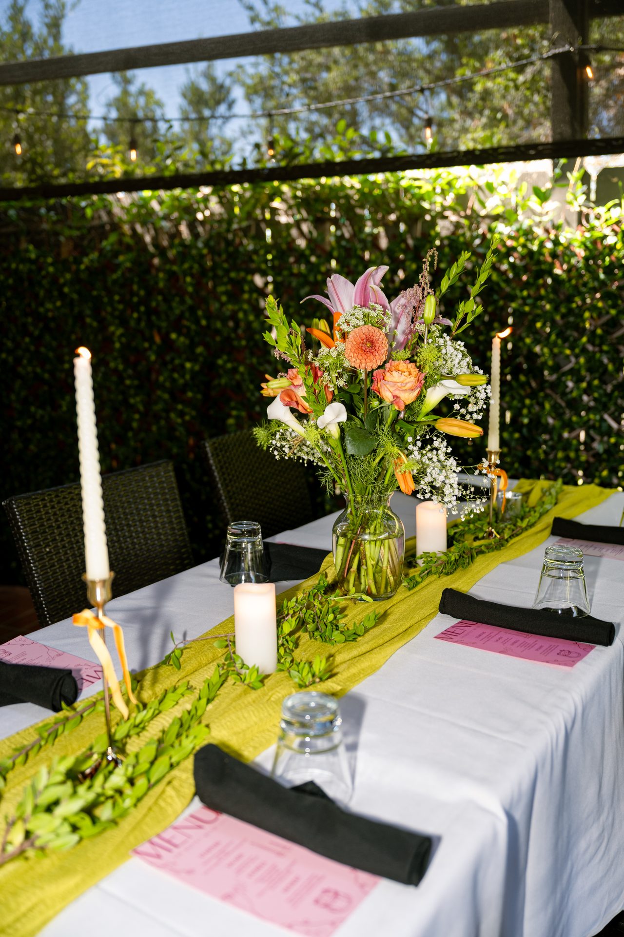 A long garden table dressed with olive linen, candles, and coral-and-pink flowers beneath patio string lights.