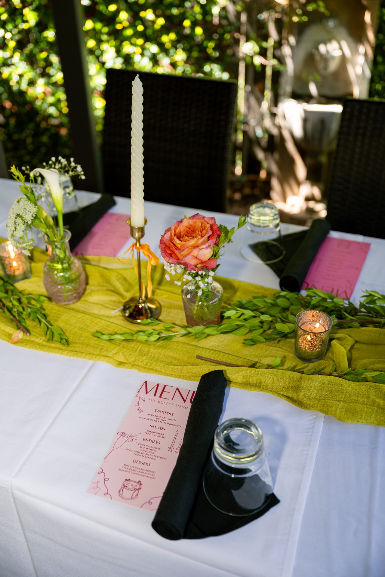 A place setting with a blush menu, black napkin, coral rose, and taper candle on an olive runner.