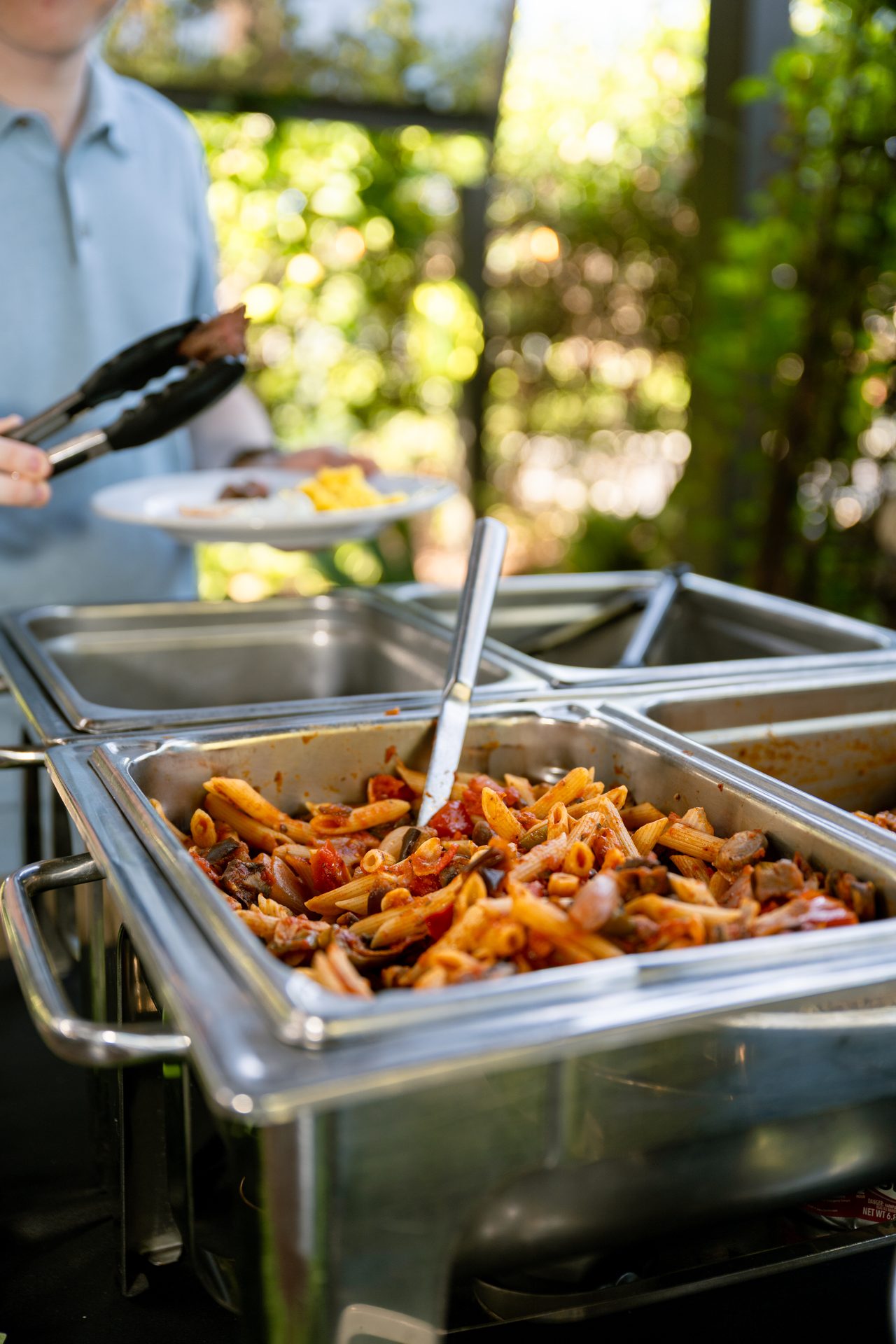 Penne pasta is served from a buffet set against the sunlit garden.
